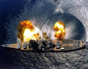  fires a full broadside of 9 x 16 in (406 mm) 50 and 6 x 5 in (127 mm) 38 guns during a target exercise. Note concussion effects on the water surface, and 16 in (406 mm) gun barrels in varying degrees of recoil.